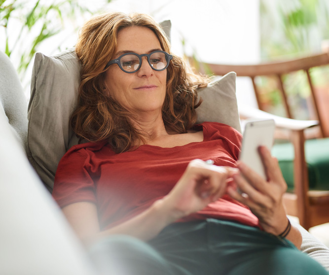 Ältere Dame liegt gemütlich auf der Couch und schaut in ihr Telefon.