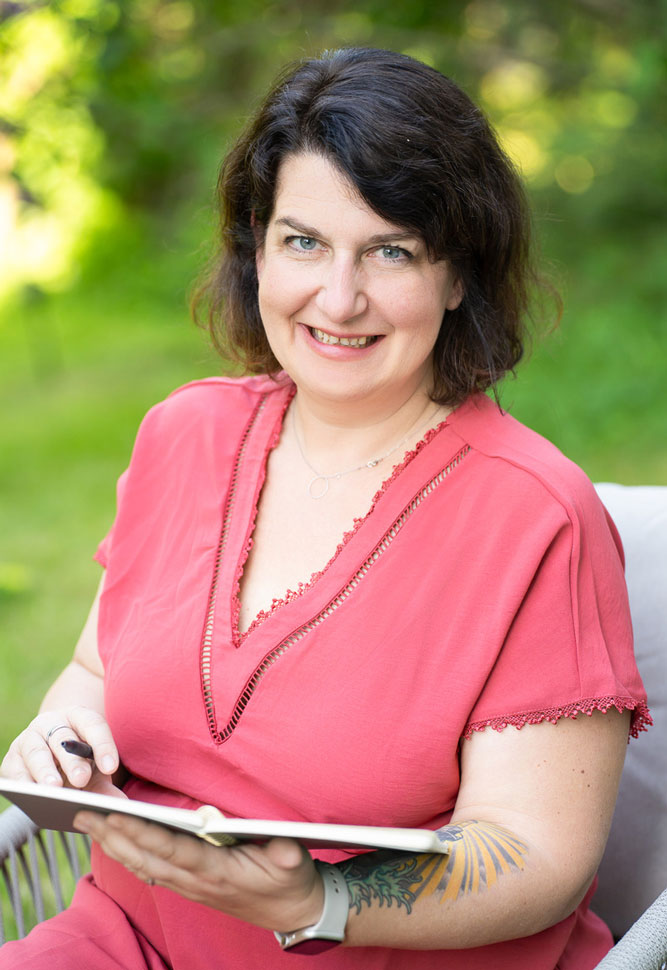 Martina Pietsch | wachsenimwechsel.at Martina Pietsch wachsenimwechsel lacht freundlich in die Kamera, mit einem Notizbuch in der Hand.